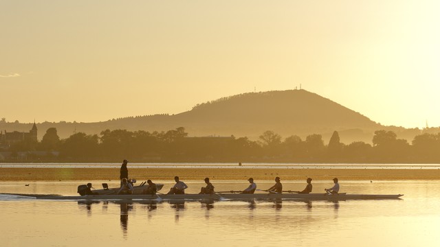 A rowing crew and a nearby motorboat are silhouetted against golden water and a distant hill.