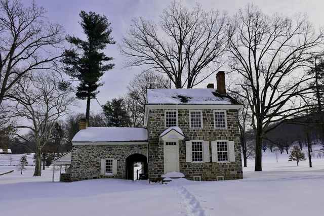 A stone house with white shutters and a lower wing, surrounded by snow and leafless trees.