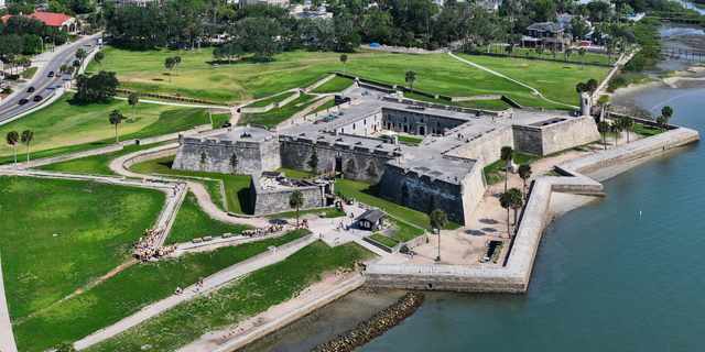An aerial view shows the stone fort’s bastions, courtyard, surrounding grass, moat and shoreline.