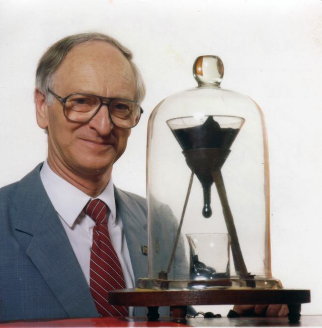 John Mainstone stands beside a glass-covered funnel of dark pitch above a beaker on a wooden base.