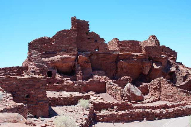 Red sandstone pueblo walls rise around a rock outcrop beneath a blue sky.