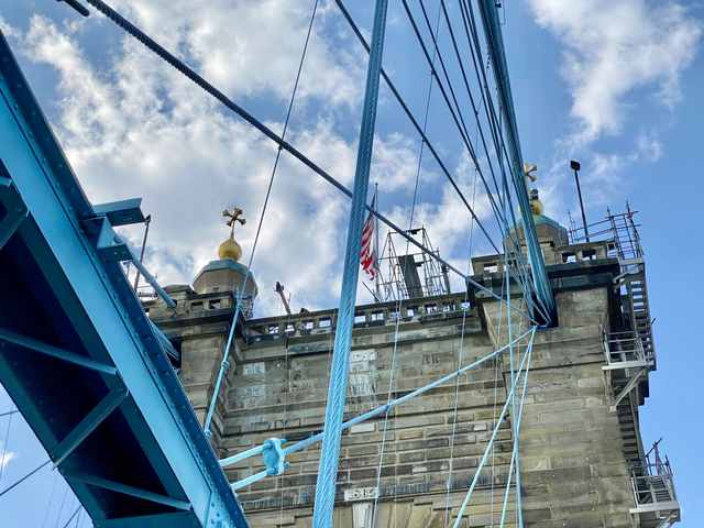 Blue bridge cables cross a close view of a stone tower with scaffolding and two small turrets.
