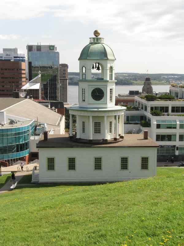 A white clock tower on the hillside, with downtown buildings and the harbour beyond; small pedestrians appear at the lower edges.