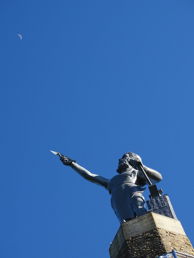 A closer low-angle view of Vulcan holding a spear point and hammer against blue sky.