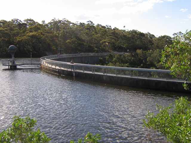 Reservoir water beside the sweeping crest of the dam, with a small intake structure and wooded banks.