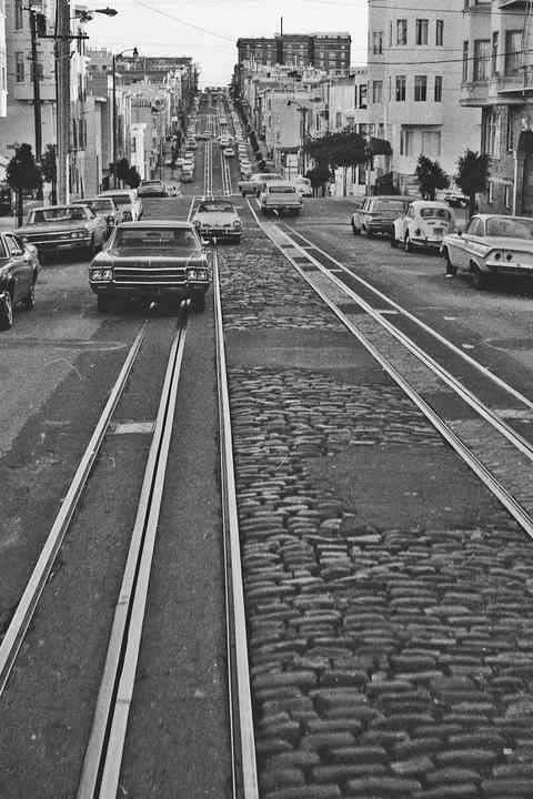 A black-and-white view along two sets of cable-car tracks and narrow slots over a hilly street lined with parked cars and buildings.