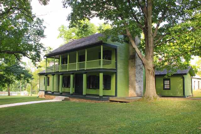 A green two-story house with dark shutters and porch railings stands beneath leafy trees.
