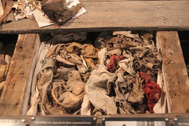 A museum display of worn fabric, footwear and fragments gathered between wooden joists, viewed through glazing.