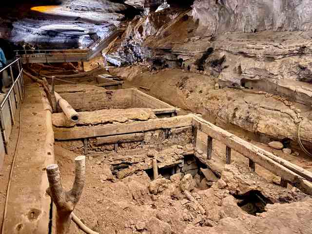 Wooden vats, scattered timbers and a hollow pipe beneath a stone cave wall, with stairs and a walkway alongside.
