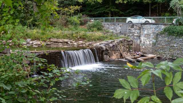 Water cascades over a low boulder dam beside a concrete channel and stone walls.