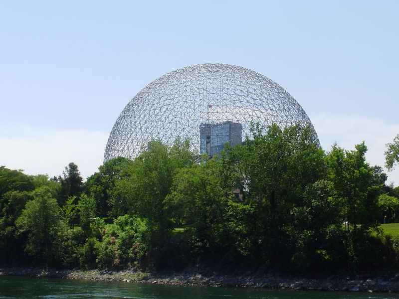 An open spherical framework rises over a leafy shoreline, with water in front and a rectangular volume visible within the dome.