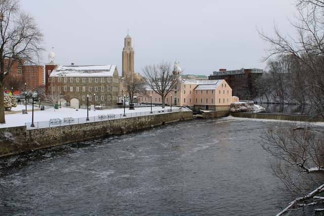 Snow covers the roofs and riverfront grounds of two mills beside a low dam, with water flowing across the foreground.