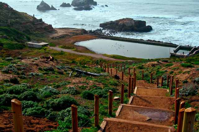 Steps descend toward water-filled basins and low walls beside the ocean, with a few distant visitors at the far right.
