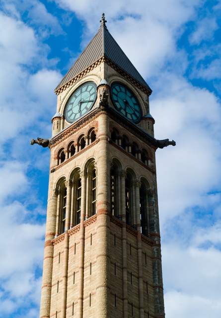 The upper part of a stone clock tower beneath a pointed roof, with projecting creatures at its corners against a blue sky.