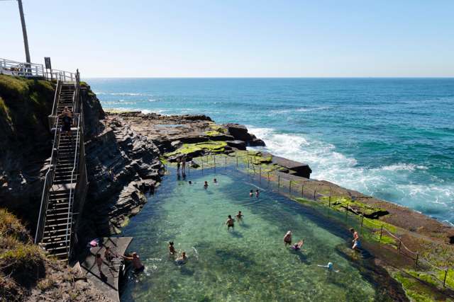 A rock-cut coastal pool beside a steep metal stair, with swimmers, a chain boundary and ocean beyond.