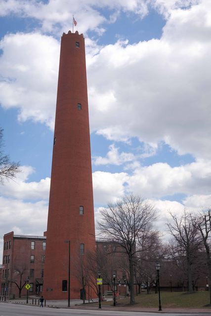 A tall tapering red-brick tower with small windows and a crenellated top beside a street.