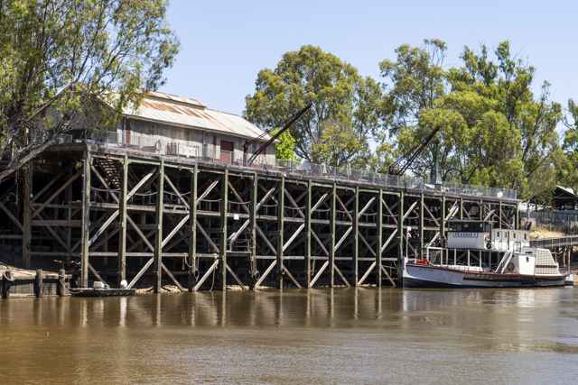 A long timber wharf with crossed braces, a shed and cranes above, and the paddle steamer Pevensey beside the riverbank.