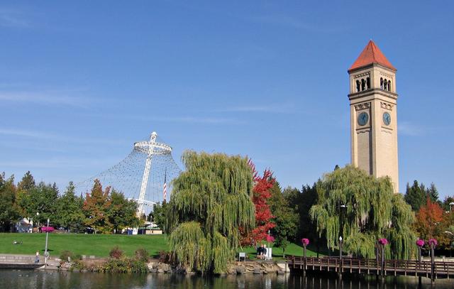 A tall clock tower stands at right, with the white cable framework of a pavilion to its left, above autumn trees, a river and a wooden footbridge.