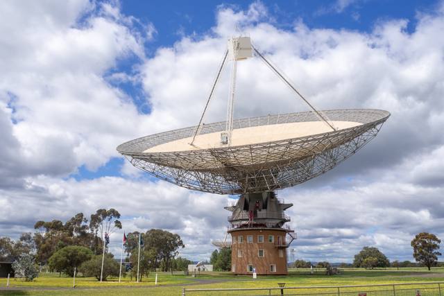 A broad white radio telescope dish and elevated focus cabin rise above a cylindrical base, green grass, trees and a foreground fence beneath clouds.