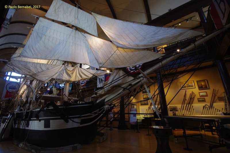 White sails and dark rigging rise above the black-and-white bow of a large ship model inside a tall museum hall.