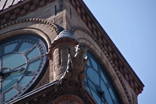 A foreshortened view of a projecting creature between two clock faces, with stone ornament and a small metal cap behind it.