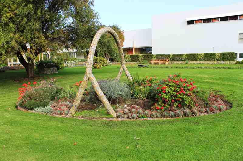 A tall stone-faced loop arches above flowers and succulents on a lawn, with a bench and factory buildings behind.