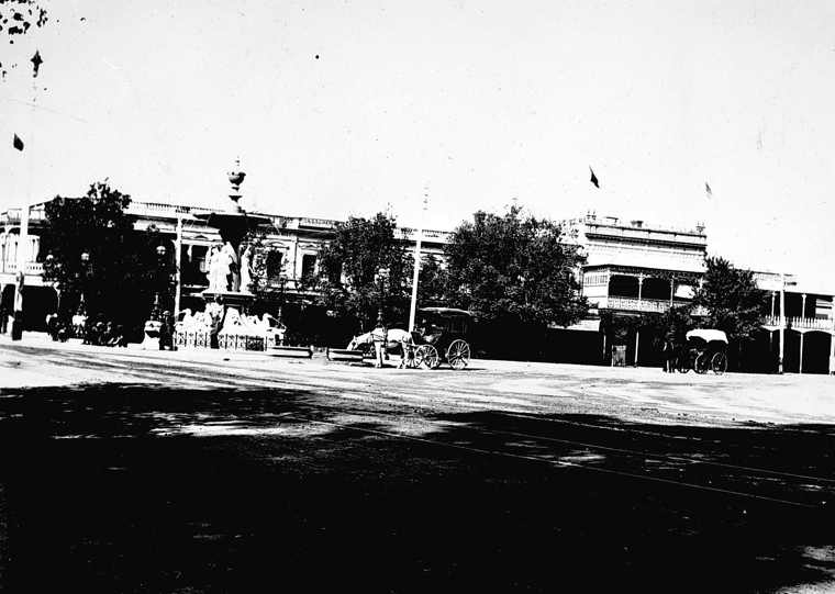 A historical black-and-white streetscape shows the fountain on the left, shopfronts and trees behind it, and horse-drawn vehicles along the road.