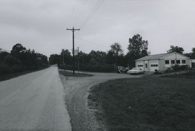 Black-and-white view of a road beside a two-bay garage, several vehicles, utility poles and trees.