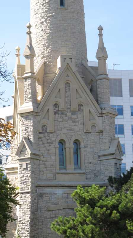 The lower stone tower and its square base have projecting buttresses, pointed stone details and paired arched windows, with leafy branches at the edges.