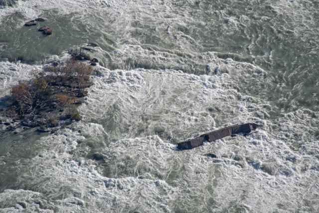 An aerial view shows the wreck amid foaming rapids, with a rocky vegetated shoal nearby.