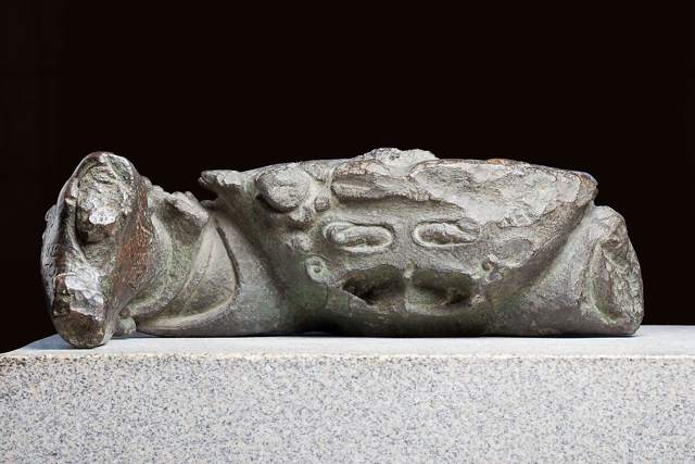 A rough-surfaced greenish bronze crab fragment on a pale stone plinth, with a raised claw at the left and faint lettering.