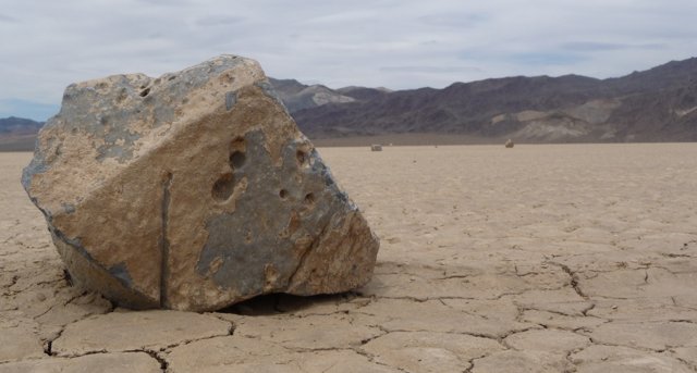 A large mottled stone on cracked pale mud, with scattered stones and mountains behind it.
