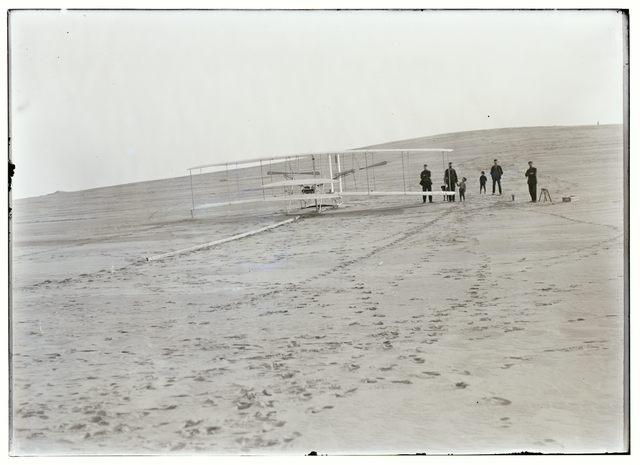 A historical photograph shows the Wright Flyer on a launching track and people beside it on a broad sandy slope.