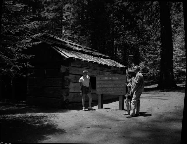 Three visitors stand beside a wooden sign in front of a log cabin with a low timber roof, in a black-and-white archival photograph.