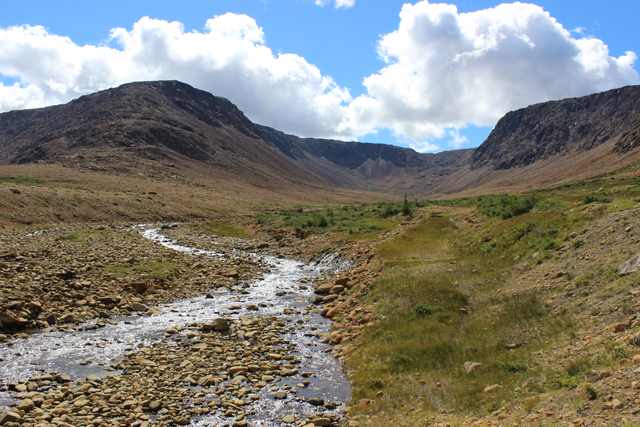 A stream curves through a stony valley below brown slopes; green plants occupy patches beside the water and along the right bank.
