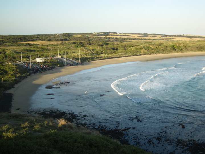 A curving sandy beach meets small waves, with viewing stands to the left and low vegetation behind the shore.