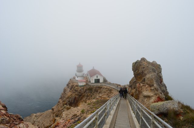 A lighthouse and adjacent building stand below a descending stairway in coastal fog.