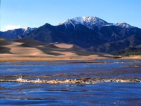 A small wave crosses shallow water, with sand dunes and snowy mountains behind.
