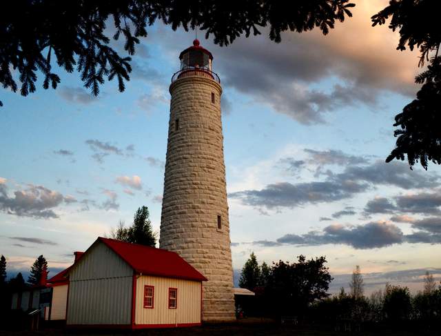 White stone lighthouse with a red lantern roof, framed by trees beside a lower red-roofed building.