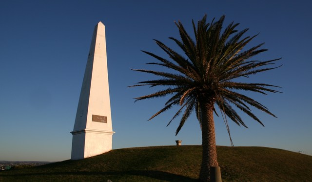 A pale, tapering obelisk with a small plaque stands beside a palm on a grassy slope under a blue sky.