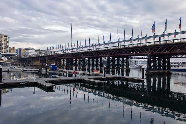 Timber trusses and piers carry a long bridge across the harbour behind floating pontoons.