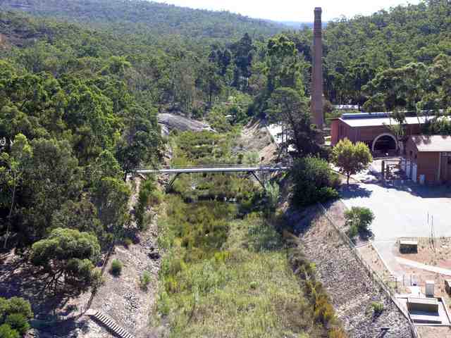 A wooded valley with a channel and footbridge, and a brick pumping-station building and tall chimney on the right.