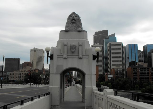 A lion sculpture sits above an arched bridge walkway, with lamps, a buffalo-head relief and city buildings beyond.
