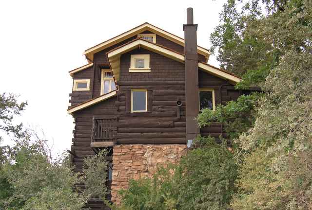 A brown wooden building with several pitched roofs, cream-trimmed windows, log and shingle walls, a stone foundation and trees.