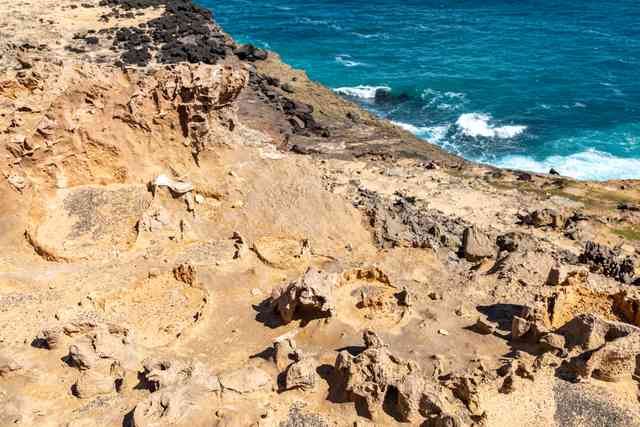 Irregular ochre formations have hollow centres and thick rims, with blue sea, surf and darker rocks behind.