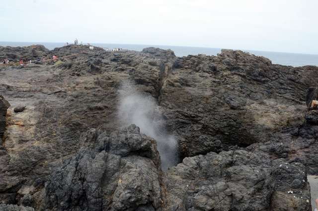 Spray rises from a coastal rock opening, with the sea and small distant visitors behind it.