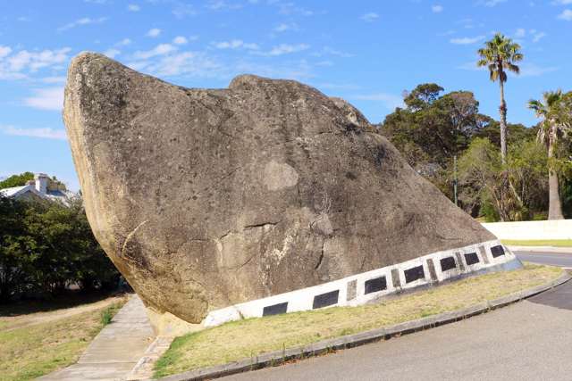The broad left side of the rock shows its dog-like profile and a white band with black rectangles beside grass and roadway.