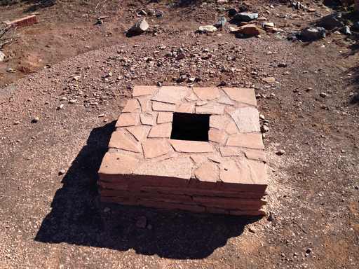 A dark square opening within a low reddish masonry surround, with rocky desert ground around it.