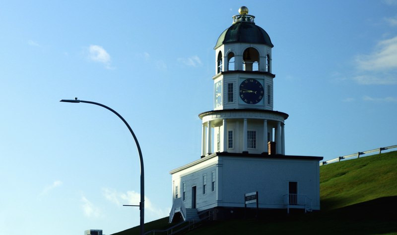 White clock tower on a grassy slope, with blue clock faces beneath a domed roof and a curved streetlamp in the foreground.