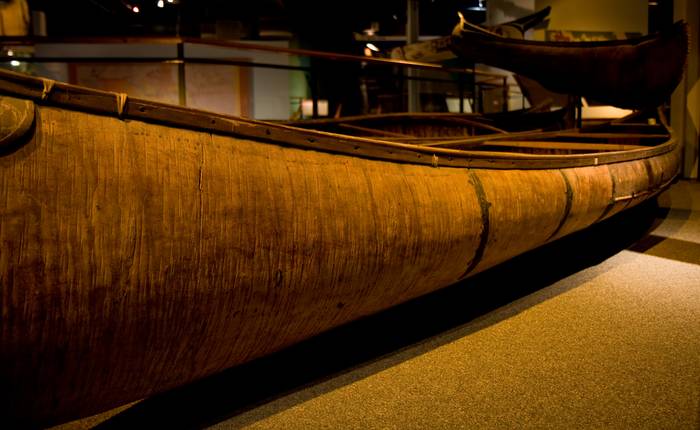 Close view of a birchbark canoe’s curved hull and crosspieces in a dimly lit museum gallery.